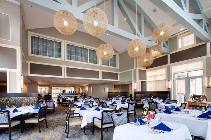 Spacious dining room with multiple tables covered in white tablecloths, each set with blue napkins, glassware, and small floral centerpieces. The room features high ceilings with large woven pendant lights hanging down, large windows allowing natural light, and a patterned carpet floor.