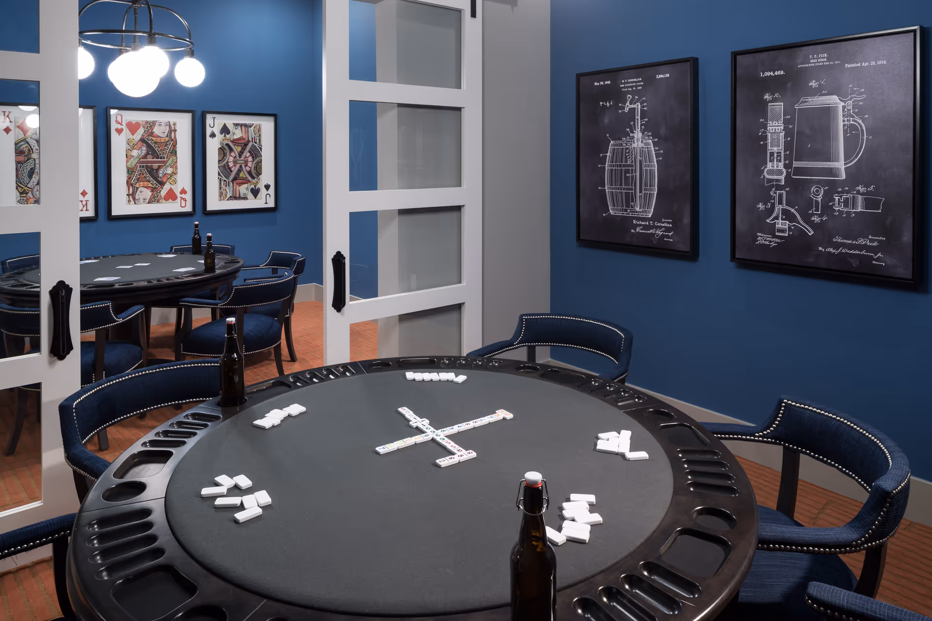 A game room with a round black table set up for dominoes, surrounded by blue upholstered chairs with nailhead trim. The walls are painted blue and decorated with framed patent-style artwork and playing card images. A glass-paned white door is partially open, revealing a reflection of the room.