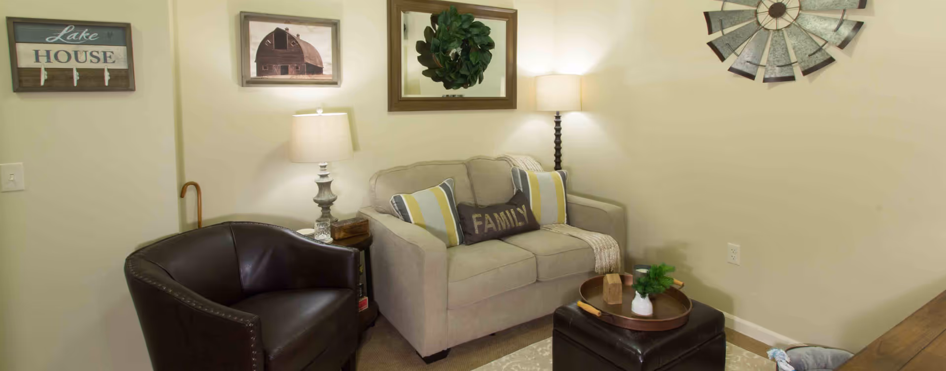 A cozy living room area featuring a beige loveseat with decorative pillows, including one that says 'FAMILY', a dark brown leather armchair, two table lamps on either side of the loveseat, a small side table, a round tray with a small plant on a dark ottoman, and wall decorations including a 'Lake House' sign, framed pictures, and a metal windmill decor.