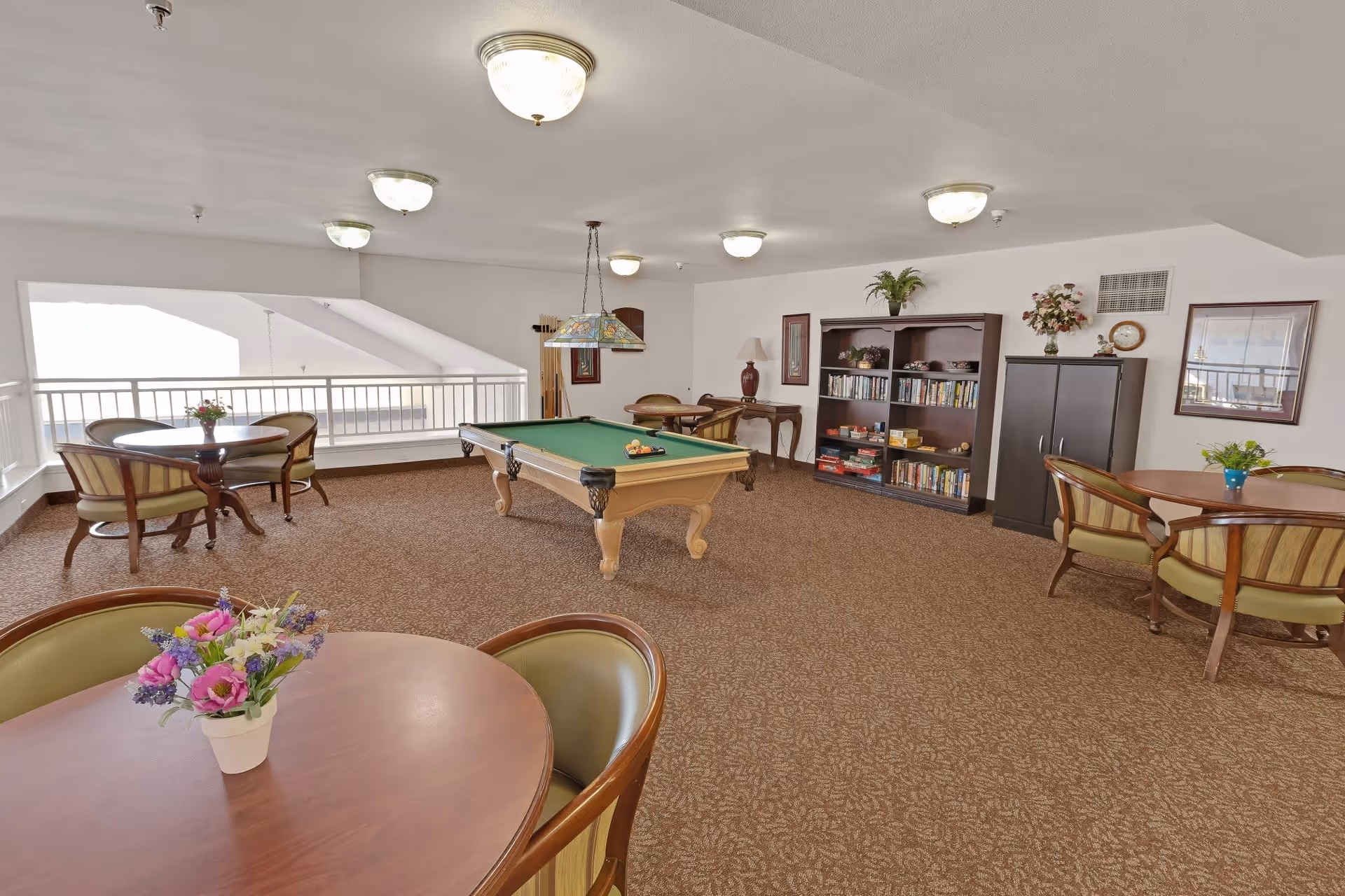 A spacious recreational room with a pool table in the center, surrounded by several round tables with chairs. The room has carpeted flooring, ceiling lights, and a bookshelf filled with books and board games. There are decorative plants and framed pictures on the walls, creating a cozy and inviting atmosphere.