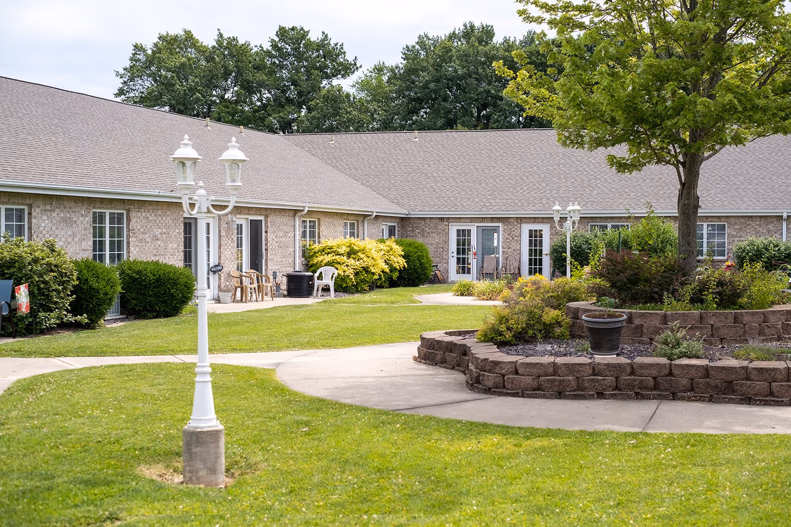 Outdoor courtyard area of a senior living facility with a well-maintained lawn, a circular raised flower bed with plants and a tree, white lampposts, and a single-story brick building with multiple doors and windows in the background.