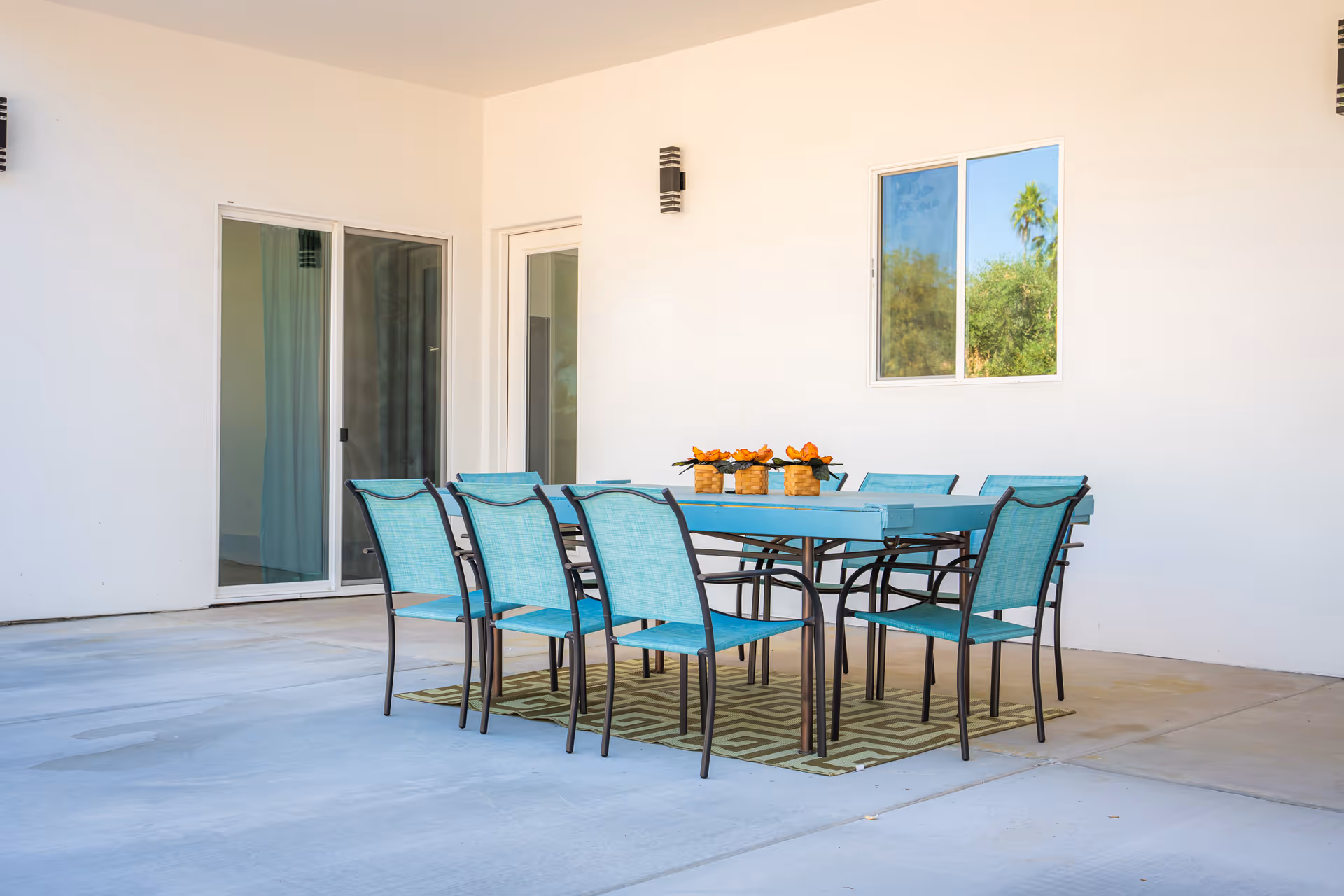 Outdoor covered patio area with a rectangular turquoise dining table surrounded by eight matching turquoise chairs. The table has three small decorative baskets with orange flowers. There is a window and a sliding glass door on the white wall behind the table.