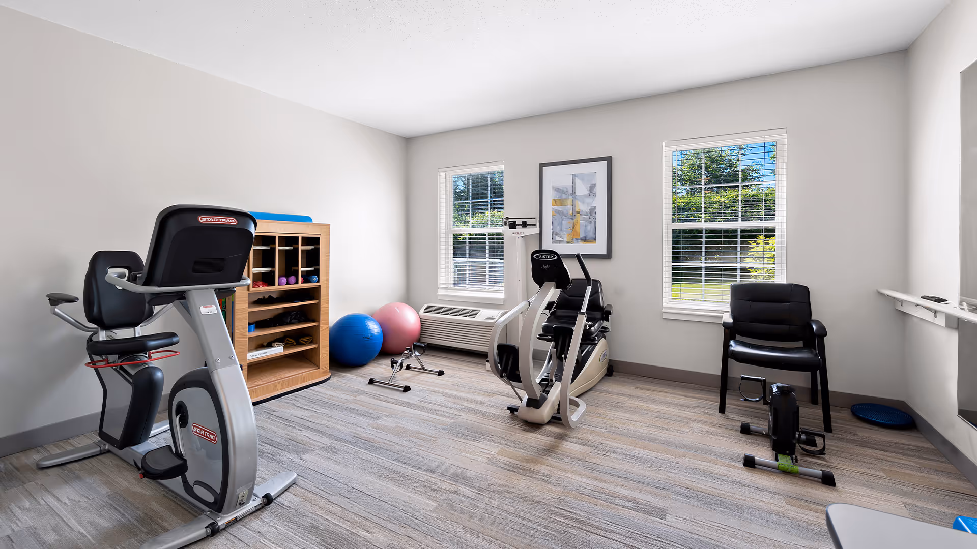 A small exercise room with two stationary exercise bikes, a wooden shelf holding small weights and exercise mats, two large exercise balls, a chair, and two windows letting in natural light.