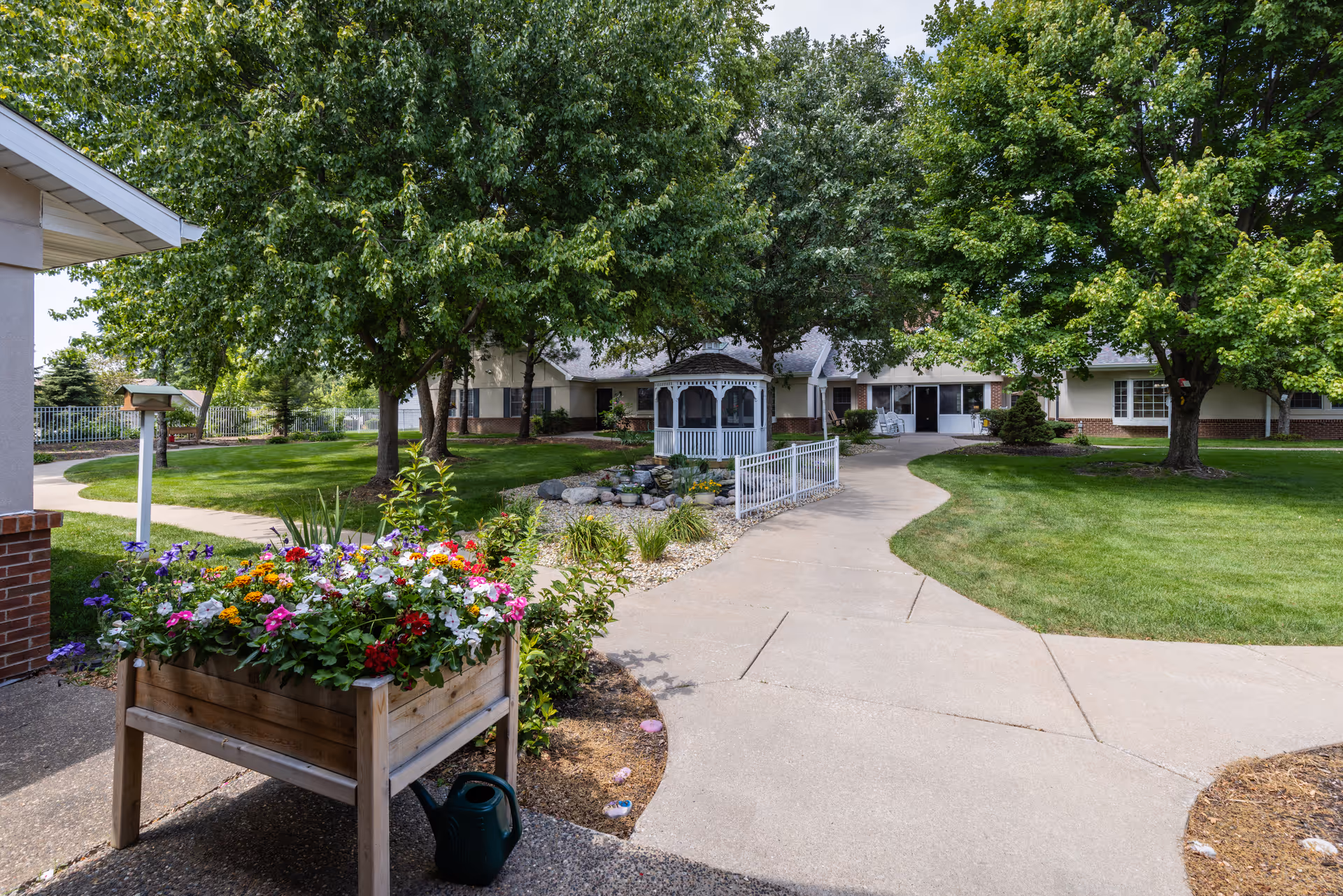 A well-maintained outdoor garden area at Country Manor Memory Care featuring a wooden planter box filled with colorful flowers, a small white gazebo surrounded by rocks and plants, a curved concrete walkway, green grass, and several large trees providing shade.