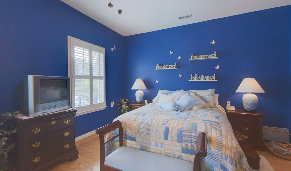 A bedroom with a bed dressed in a patchwork quilt against a blue accent wall, two bedside tables with lamps, a dresser with a TV, and a window with shutters.