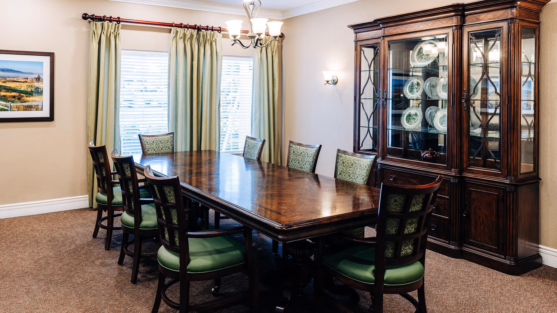 A formal dining room with a large wooden dining table surrounded by eight chairs with green upholstered seats and backs. Behind the table is a wooden china cabinet displaying decorative plates. The room has beige walls, a window with green curtains, a framed landscape picture on the wall, and a chandelier above the table.