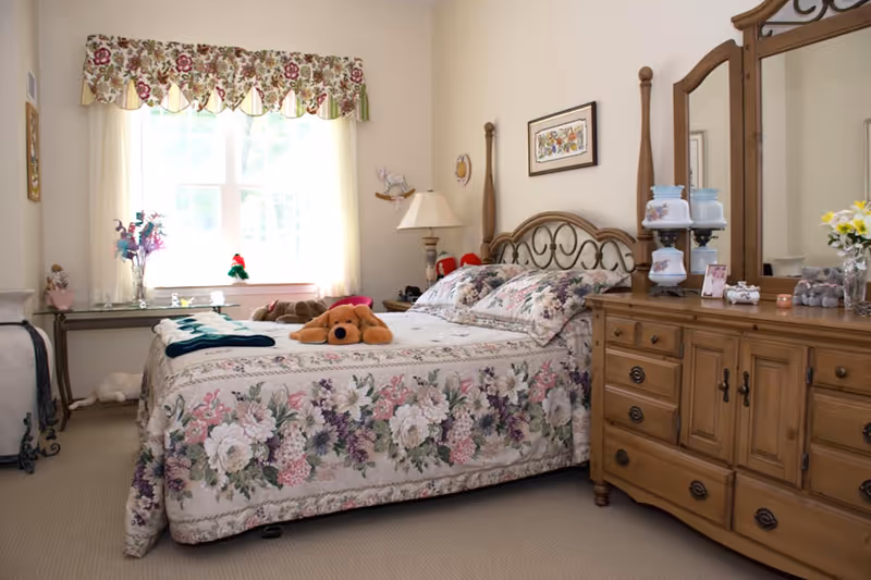 A cozy bedroom featuring a floral bedspread with a stuffed dog on the bed, a wooden dresser with mirror, and a window with a floral valance.