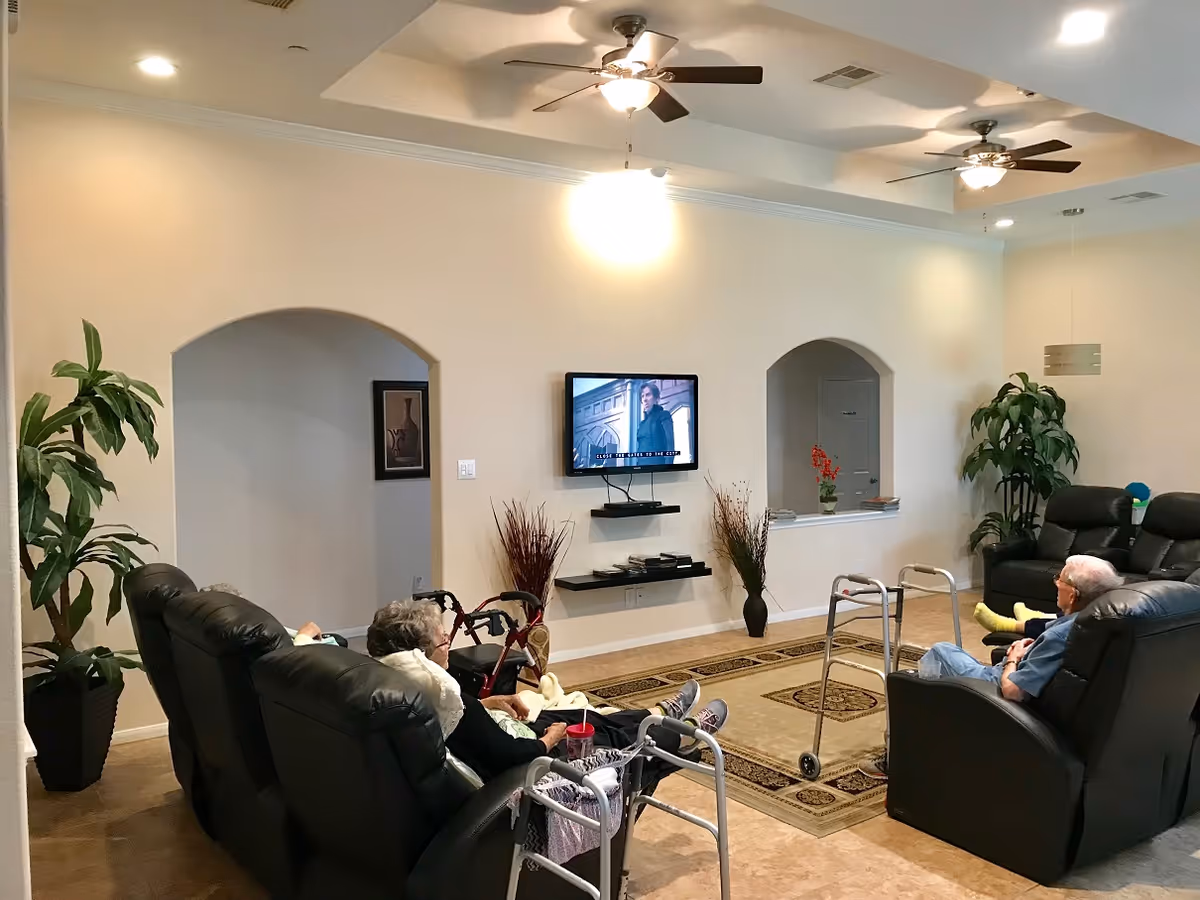 A living room in an assisted living facility with two elderly people seated in black recliners facing a wall-mounted TV. The room has beige walls, two ceiling fans with lights, potted plants, a patterned area rug, and walking aids near the seated individuals.