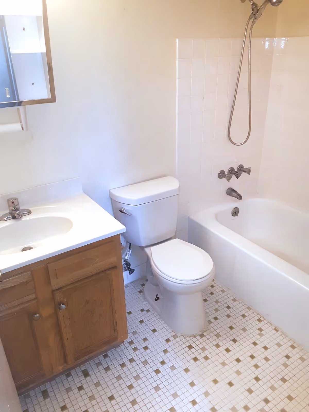 A small bathroom featuring a white toilet, a bathtub with a showerhead and faucet, a sink with a wooden cabinet below, and a tiled floor with a white and beige pattern.