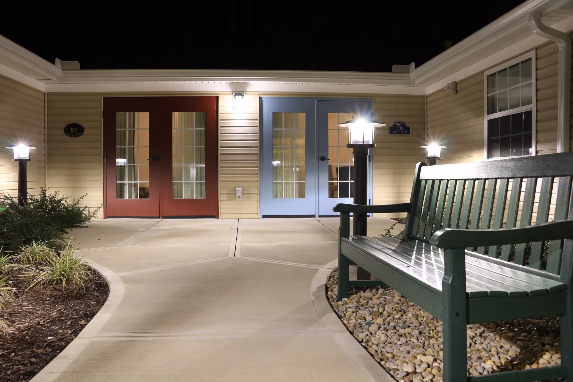 Night view of an outdoor courtyard area at a senior living facility with two sets of glass-paneled doors, one painted red and the other blue. The area is well-lit with lamp posts and a wall light above the doors. A green bench sits on a bed of small rocks to the right, and there are landscaped plants on the left side of the concrete pathway.
