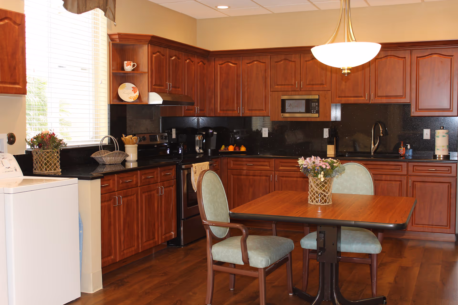 A kitchen area with wooden cabinets, a black countertop, and black appliances including a stove and microwave. There is a wooden table with two green cushioned chairs and a flower arrangement in a basket on the table. A washing machine is visible on the left side near a window with blinds.