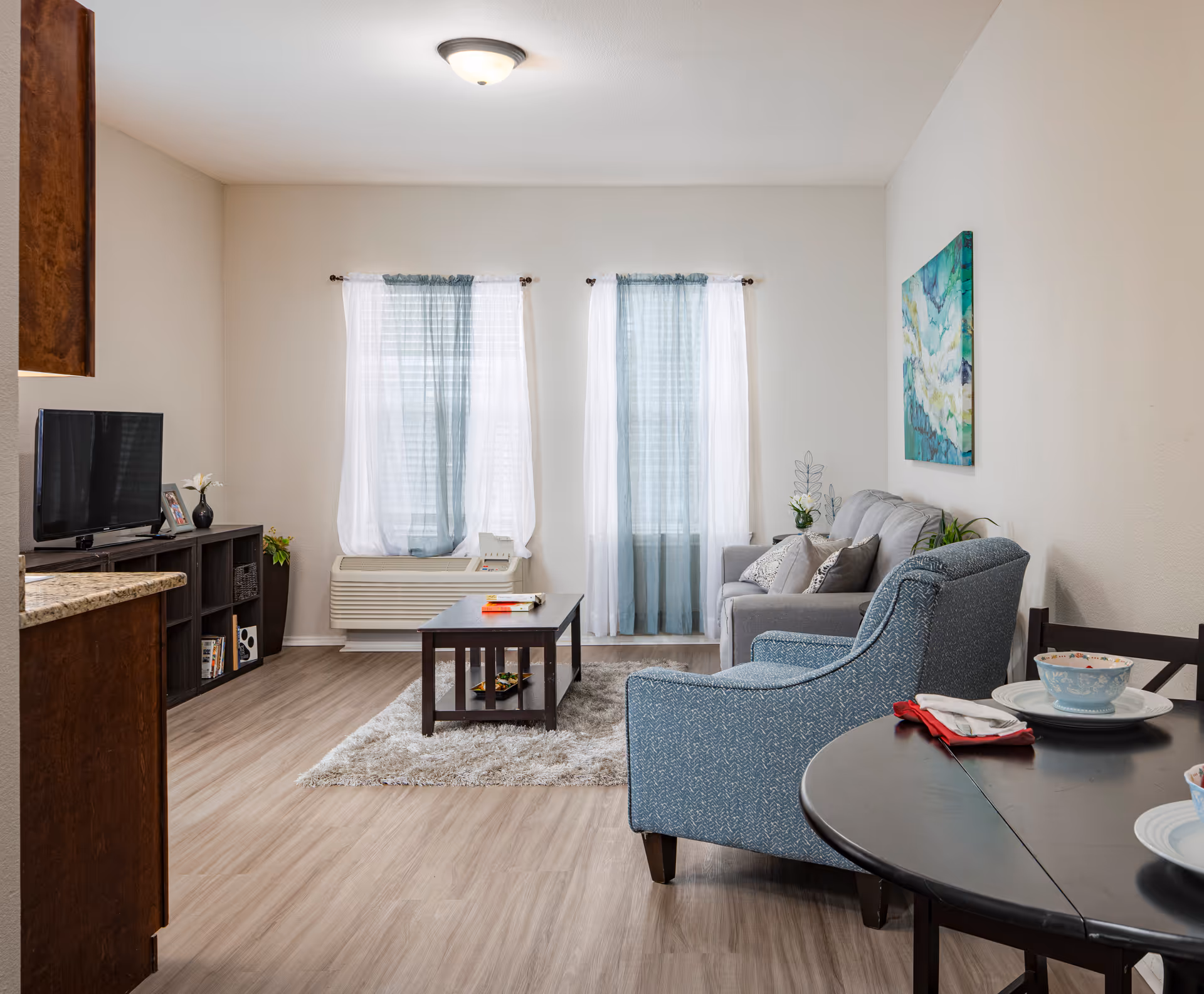 A bright and cozy living room with light wood flooring, two large windows with sheer white and blue curtains, a gray sofa with cushions, a blue armchair, a dark wood coffee table on a light gray rug, a TV on a dark wood stand, and a round dining table set with bowls and plates.