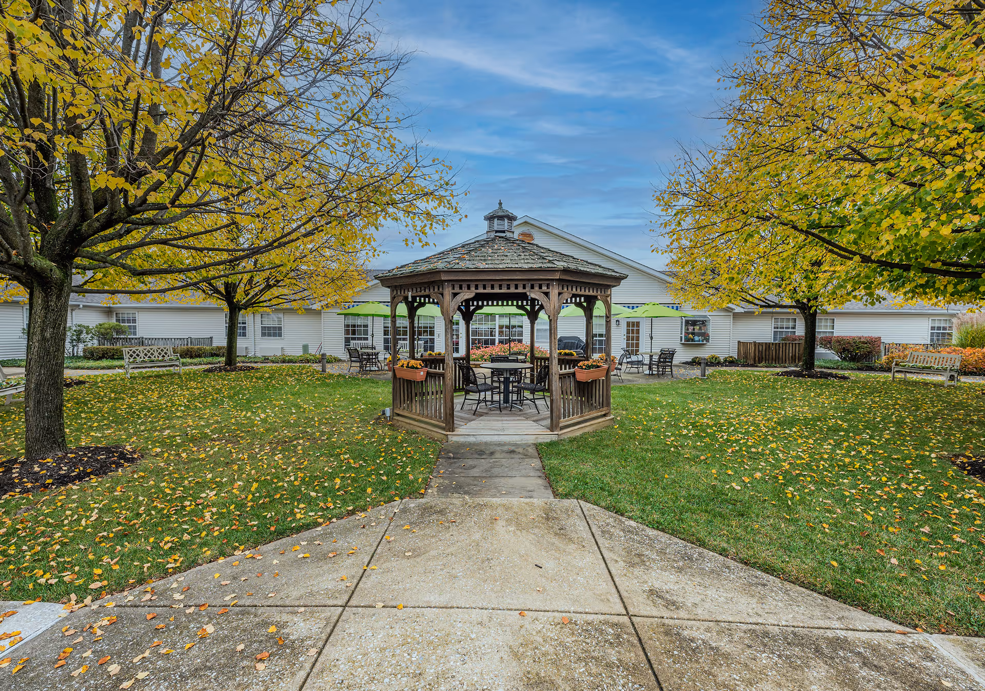 Outdoor courtyard area with a wooden gazebo in the center, surrounded by green grass and trees with yellow autumn leaves. There are benches and tables with green umbrellas around the gazebo, and a white building in the background under a partly cloudy blue sky.