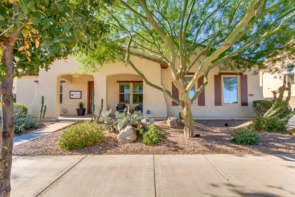 Front exterior view of a single-story building with beige stucco walls, brown window shutters, and a covered porch with two chairs. The landscaping includes desert plants, cacti, rocks, and a tree with green foliage. A sign near the entrance reads 'Alder Leaf'.