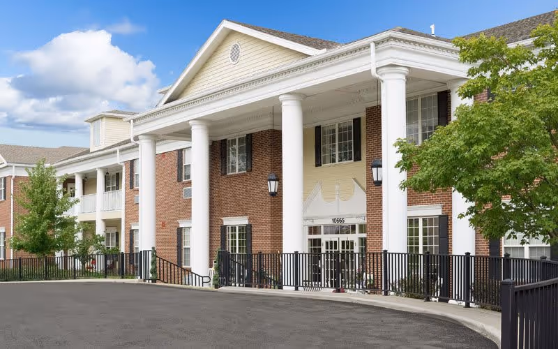 Exterior view of a senior living facility building with large white columns, brick walls, multiple windows, and a black metal fence in front. There is a tree on the right side and a clear blue sky with some clouds above.
