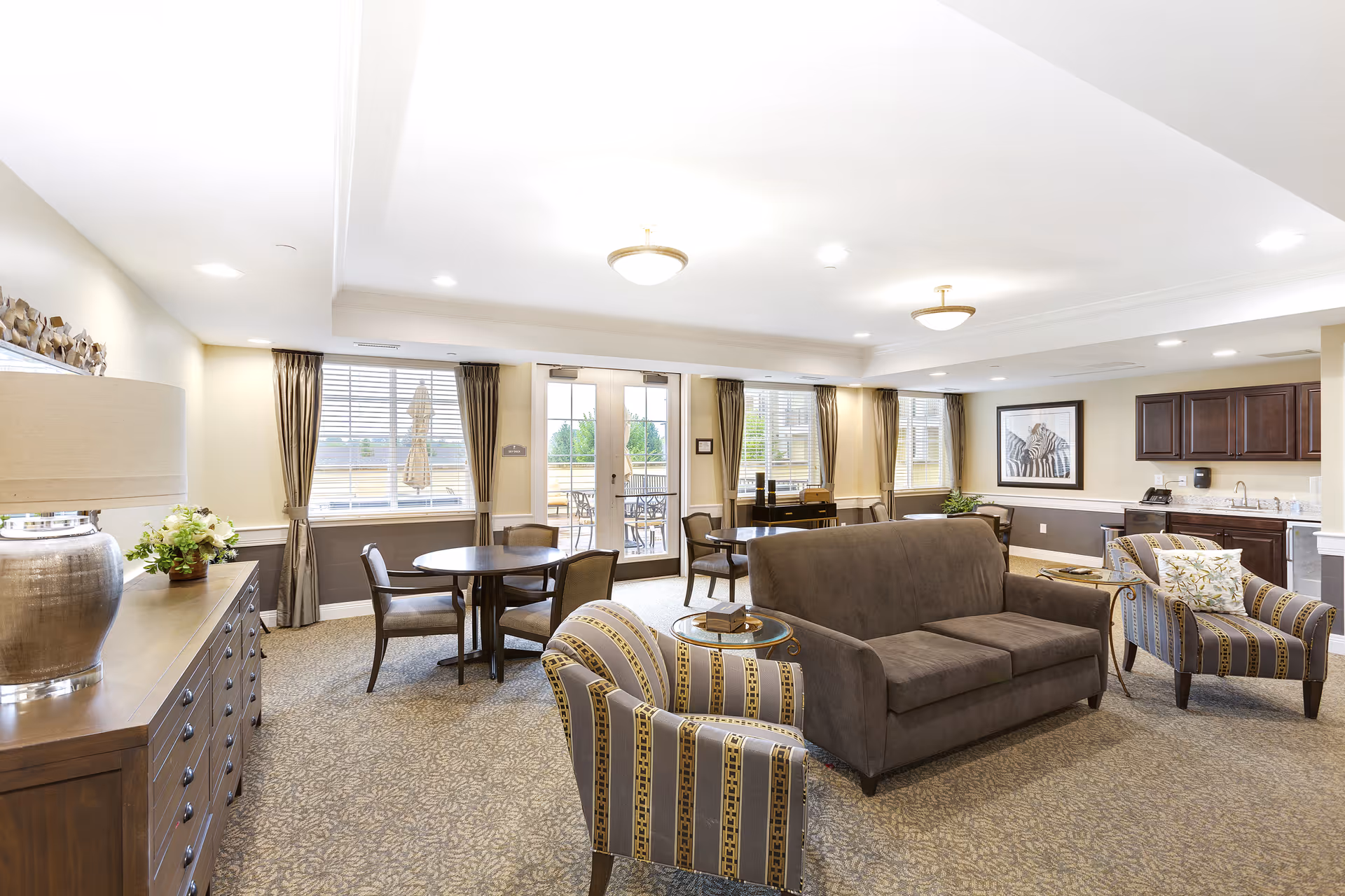 A bright and spacious living room area in a senior living facility with a brown sofa, two patterned armchairs, a round wooden table with chairs, large windows with curtains, and a kitchenette with dark wood cabinets. The room is carpeted and decorated with a lamp, plants, and framed artwork on the wall.