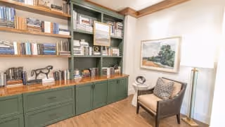 A cozy reading nook featuring a green built-in bookshelf filled with books and decorative items, a wooden chair with a patterned cushion, a small side table with a decorative bowl, a floor lamp, and a framed landscape painting on the wall.