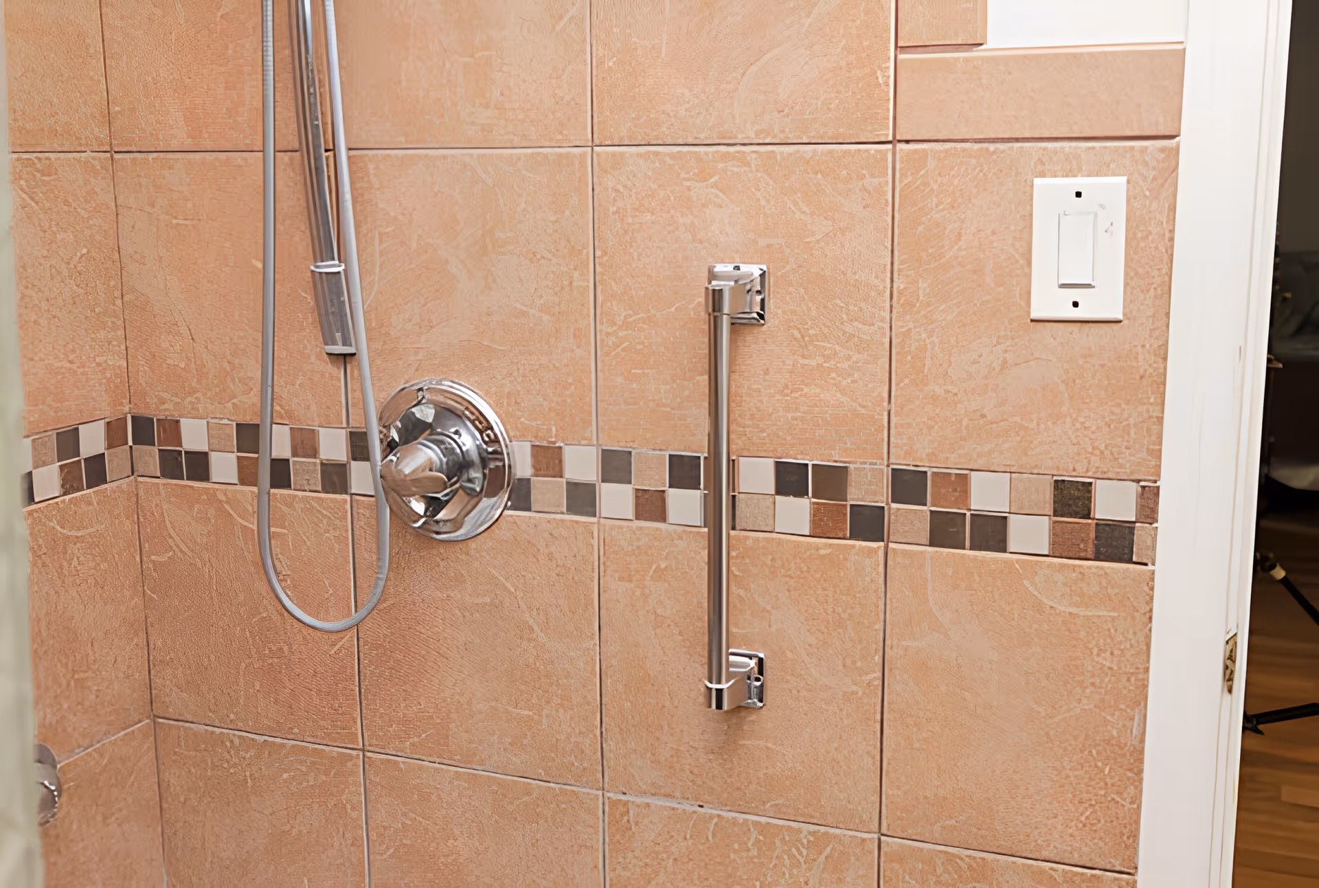 Close-up view of a shower wall with beige tiles and a decorative horizontal strip of small square tiles in various shades of brown and beige. The shower features a chrome shower handle and a vertical grab bar. A white light switch is visible on the adjacent wall near the door frame.