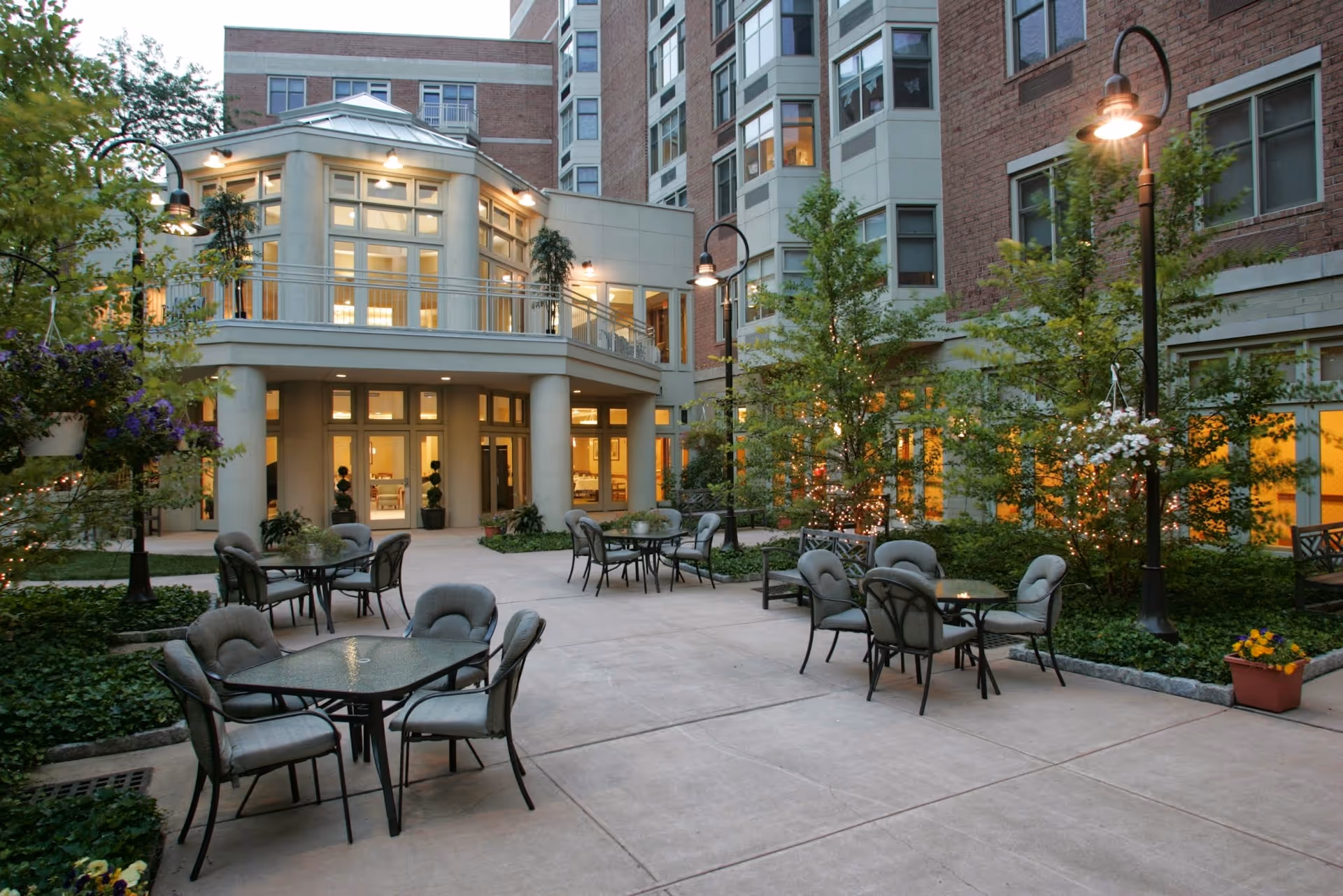 Outdoor courtyard area of a senior living facility with several glass-top tables and cushioned chairs arranged on a concrete patio. The courtyard is surrounded by greenery, small trees with string lights, hanging flower baskets, and tall street lamps. The multi-story building in the background has large windows and warm interior lighting visible through them.