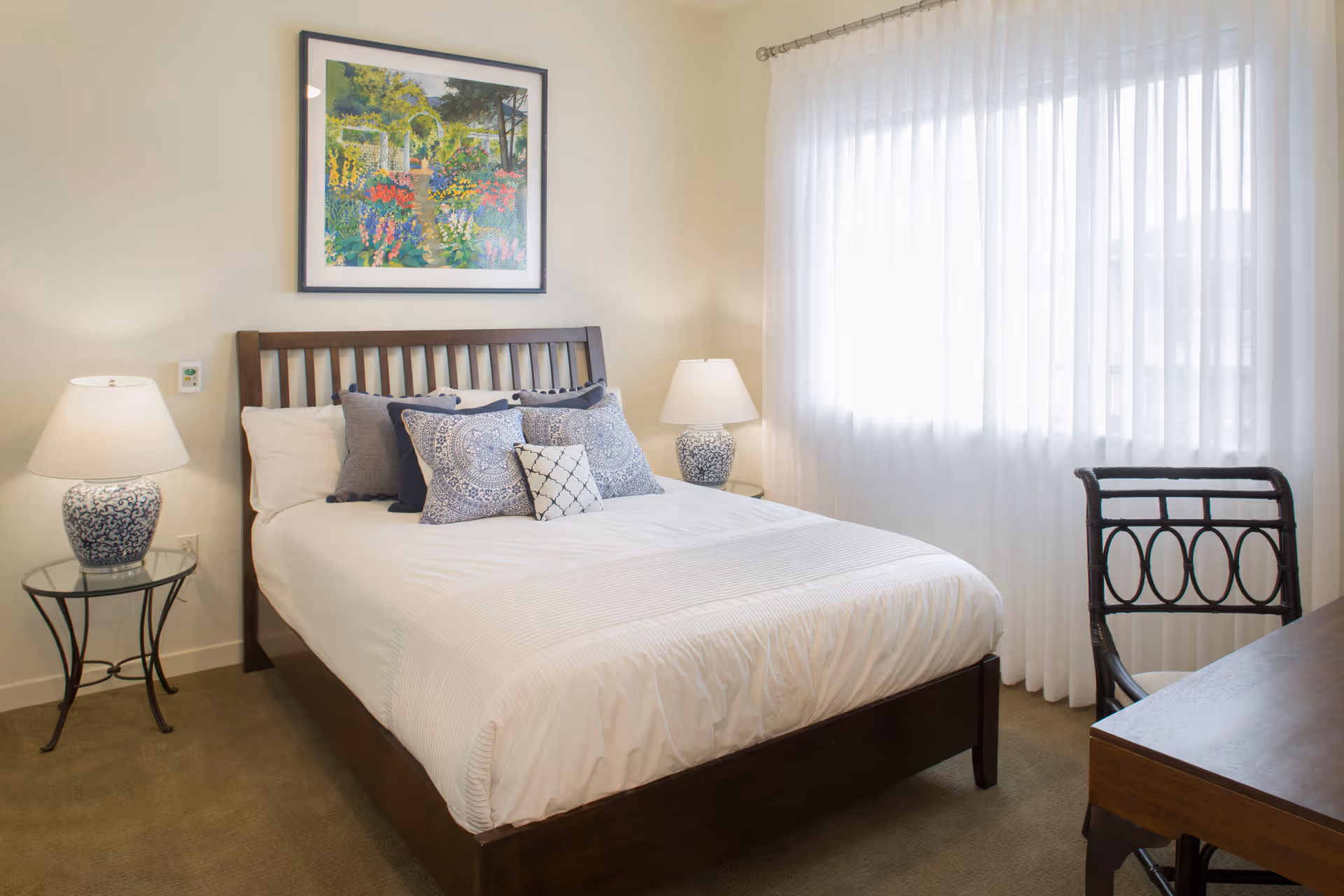 A neatly made bed with white linens and multiple decorative pillows in shades of blue and white. The bed has a dark wooden frame and is flanked by two glass-top nightstands, each with a blue and white patterned lamp. A framed colorful painting of a garden scene hangs above the bed. To the right, sheer white curtains cover a window, and a dark wooden chair with a decorative backrest is positioned next to a matching wooden desk.