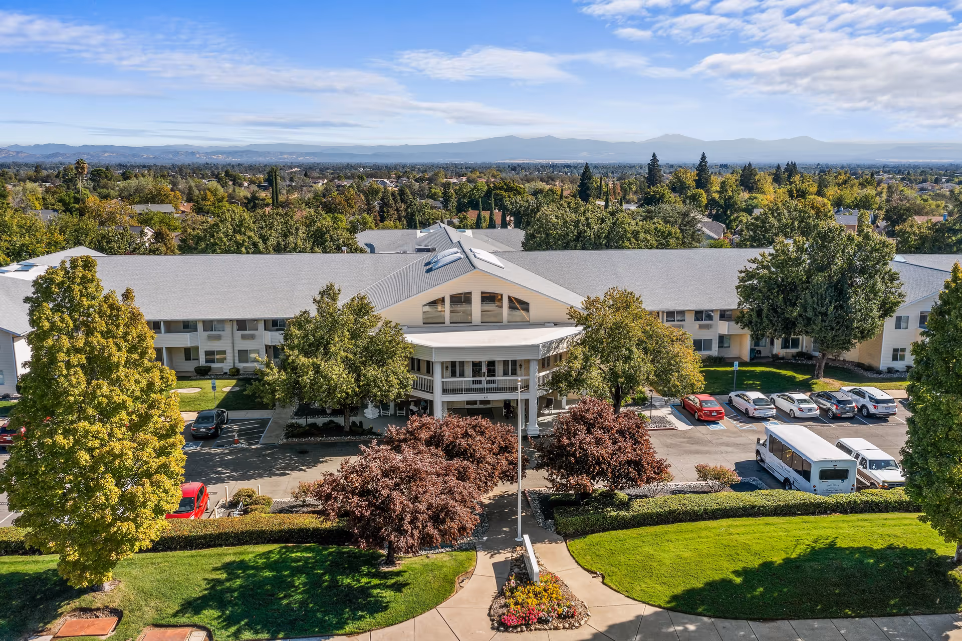 Aerial view of Hilltop Estates, a senior living community building surrounded by trees and greenery. The building has a large entrance with a covered porch, parking spaces with cars and a shuttle bus, and a well-maintained lawn with flower beds and shrubs in the foreground. Mountains and a partly cloudy sky are visible in the background.
