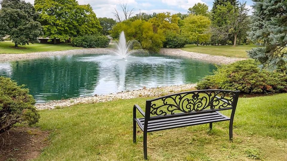 A decorative black metal bench on a grassy area facing a small pond with a water fountain in the center. The pond is surrounded by rocks and lush green trees and bushes under a partly cloudy sky.