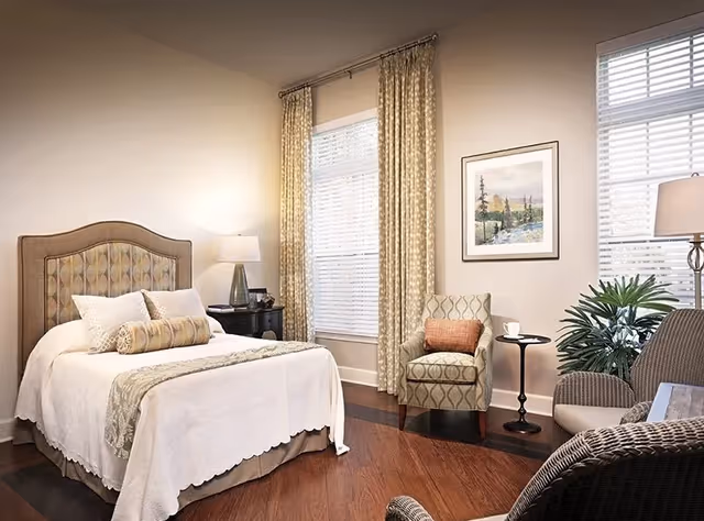 A cozy bedroom with a neatly made bed featuring a patterned headboard and decorative pillows. There is a nightstand with a lamp beside the bed, two windows with patterned curtains, an armchair with a cushion, a small round side table with a cup, a floor lamp, and a potted plant. A framed landscape painting hangs on the wall.