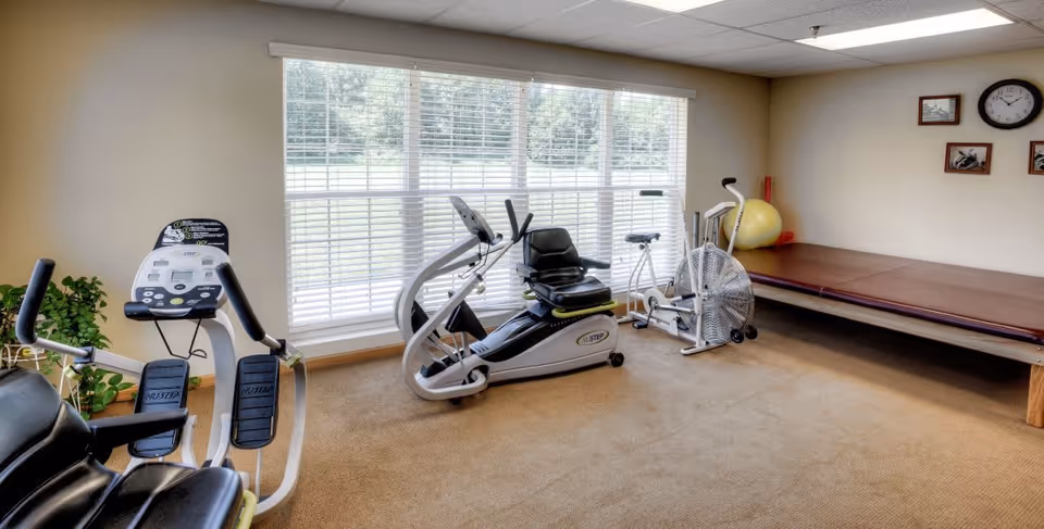 A small exercise room with two stationary exercise bikes, a large window with white blinds letting in natural light, a brown padded bench along one wall, a yellow exercise ball, and a few framed pictures and a clock on the wall.