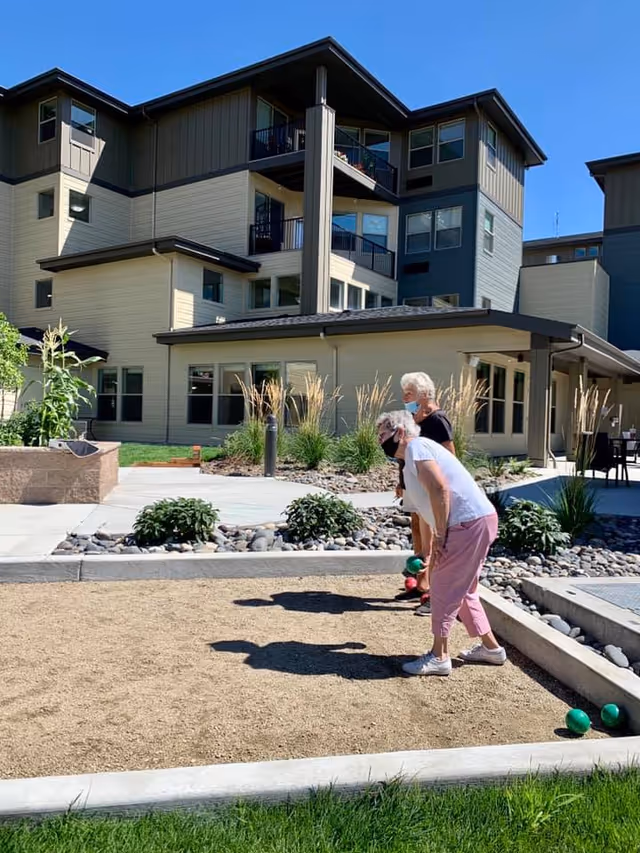 Two elderly women wearing face masks are playing bocce ball on a sandy court outside a multi-story residential building under a clear blue sky. The area is landscaped with plants and rocks, and there is a paved walkway nearby.