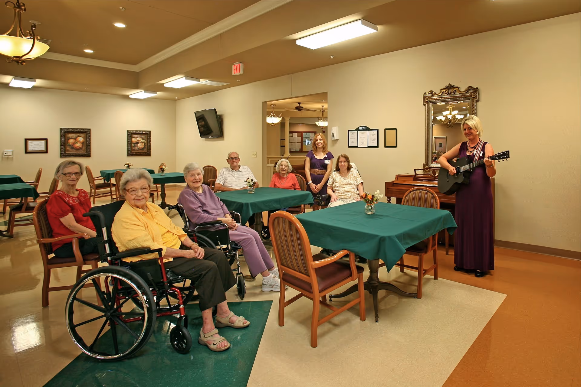 A group of elderly people sitting around tables with green tablecloths in a common room. A woman in a purple dress is standing and playing a guitar near a piano and a large mirror on the wall. The room is well-lit with ceiling lights and decorated with framed pictures and a menu on the wall.
