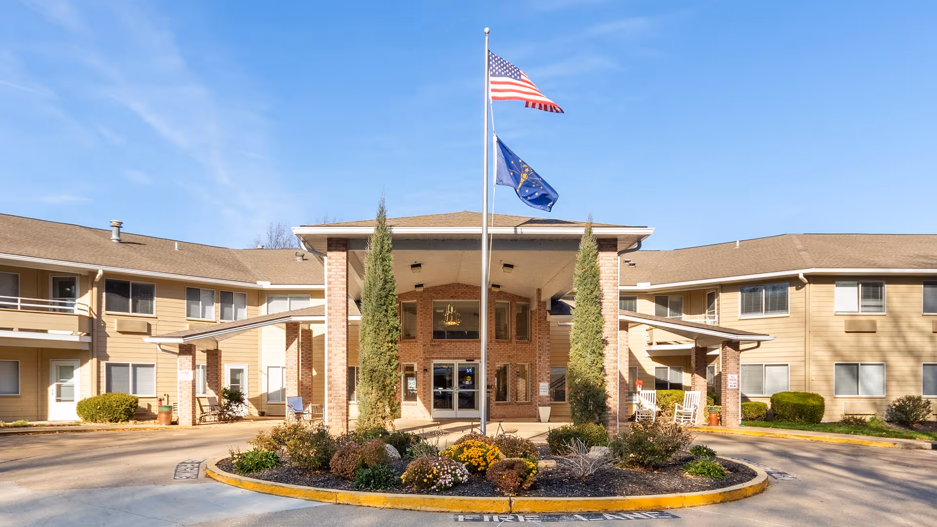 Front exterior view of a senior living facility building with a covered entrance, two tall evergreen trees, a circular flower bed with various plants and flowers, and two flagpoles displaying the American flag and a state flag against a clear blue sky.