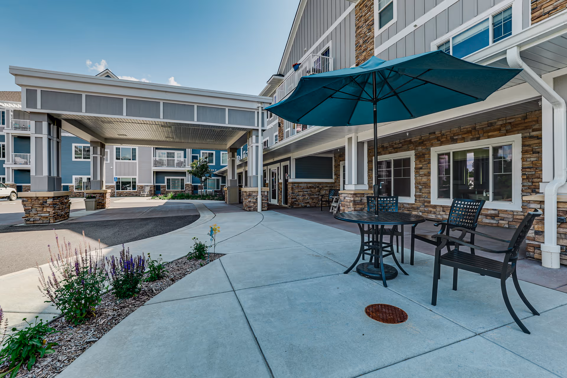 Outdoor seating area at a senior living facility with a round table, two chairs, and a large blue umbrella. The building exterior features stone and siding, with windows and a covered driveway entrance. There are small flower beds along the sidewalk.