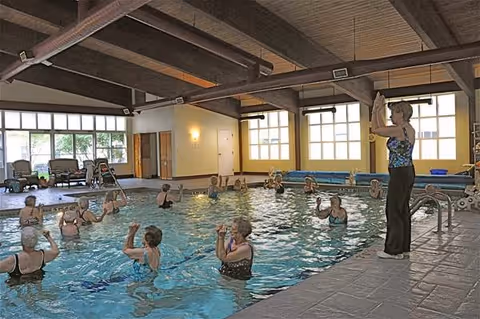 Indoor swimming pool with a group of elderly people participating in a water exercise class led by an instructor standing at the poolside. The room has large windows, exposed wooden beams on the ceiling, and poolside seating.