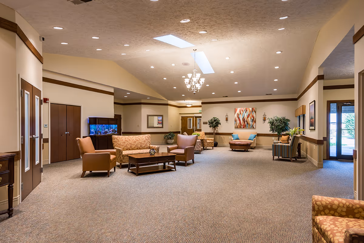 A spacious and well-lit common area in a senior living facility featuring multiple seating arrangements with armchairs, sofas, and a coffee table. The room has beige walls with wooden trim, a carpeted floor, recessed lighting, a chandelier, skylights, and decorative plants. There is a large fish tank against one wall and artwork hanging on the walls. Double doors and large windows provide natural light and access to the outside.