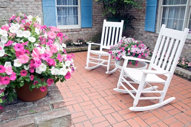 A cozy outdoor patio area with two white wooden rocking chairs and a small round glass-top table between them. The patio is paved with red bricks and decorated with large pots of pink and white flowers. The background shows a brick wall with blue window shutters and a window with white curtains.
