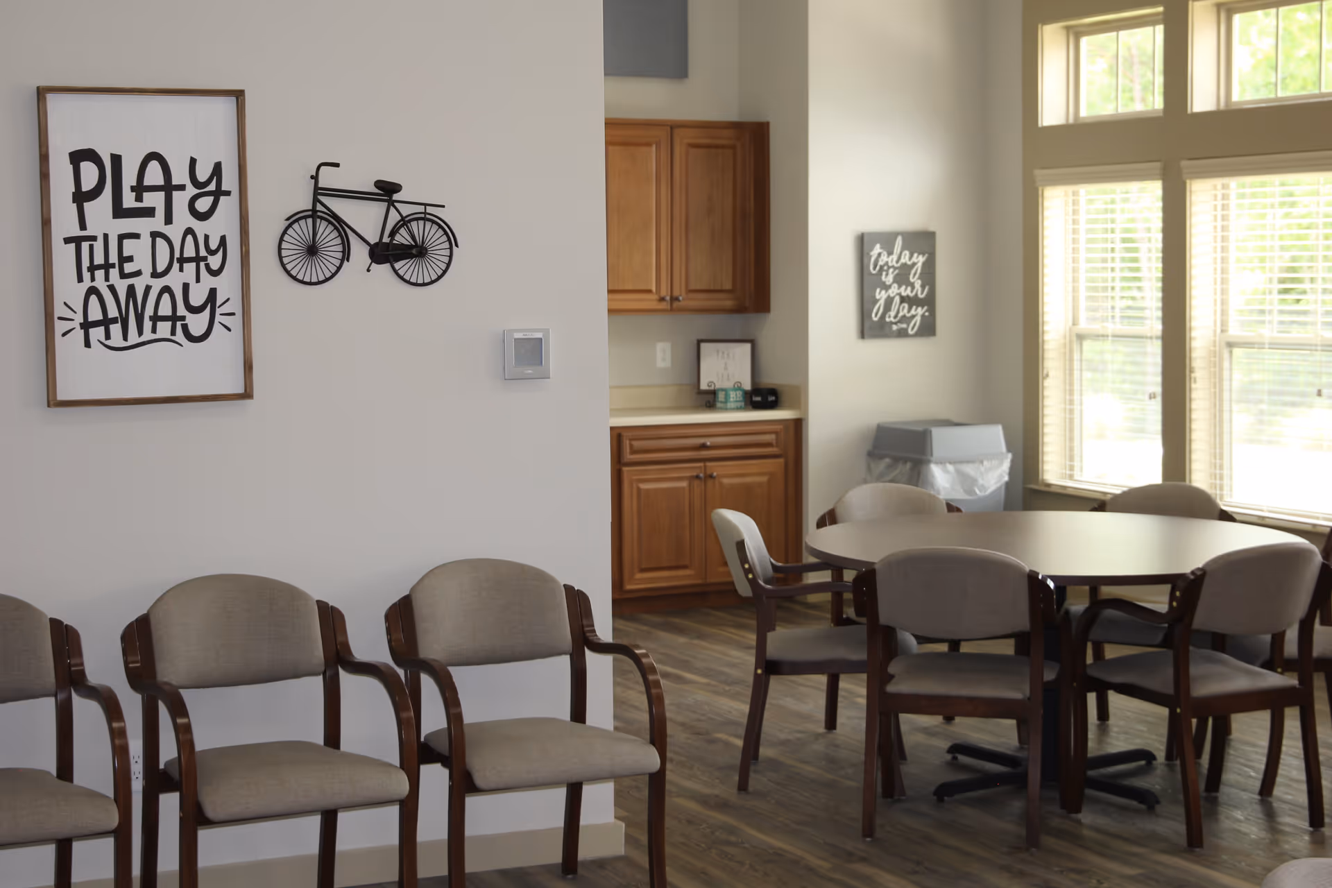 A bright room with several beige cushioned chairs arranged along a wall and around a round table. The wall features a decorative metal bicycle and a framed sign that reads 'PLAY THE DAY AWAY.' There are wooden cabinets and a countertop in the background, along with large windows letting in natural light. Another framed sign on the far wall reads 'today is your day.'