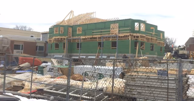 Construction site of a building with green sheathing and wooden framing on the upper floor, surrounded by construction materials and equipment, with a chain-link fence in the foreground.