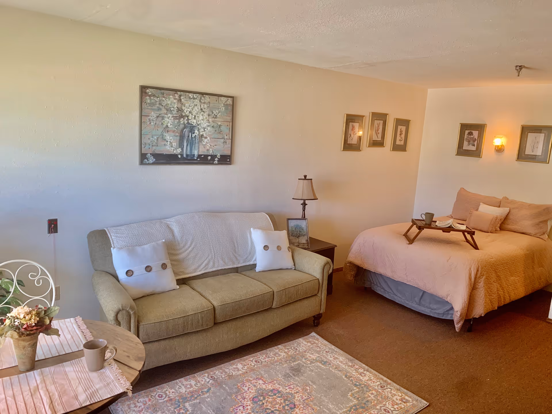 Studio-style living area with a green couch, small table and rug, and a bed with pink bedding, decorated with wall art and lamps.