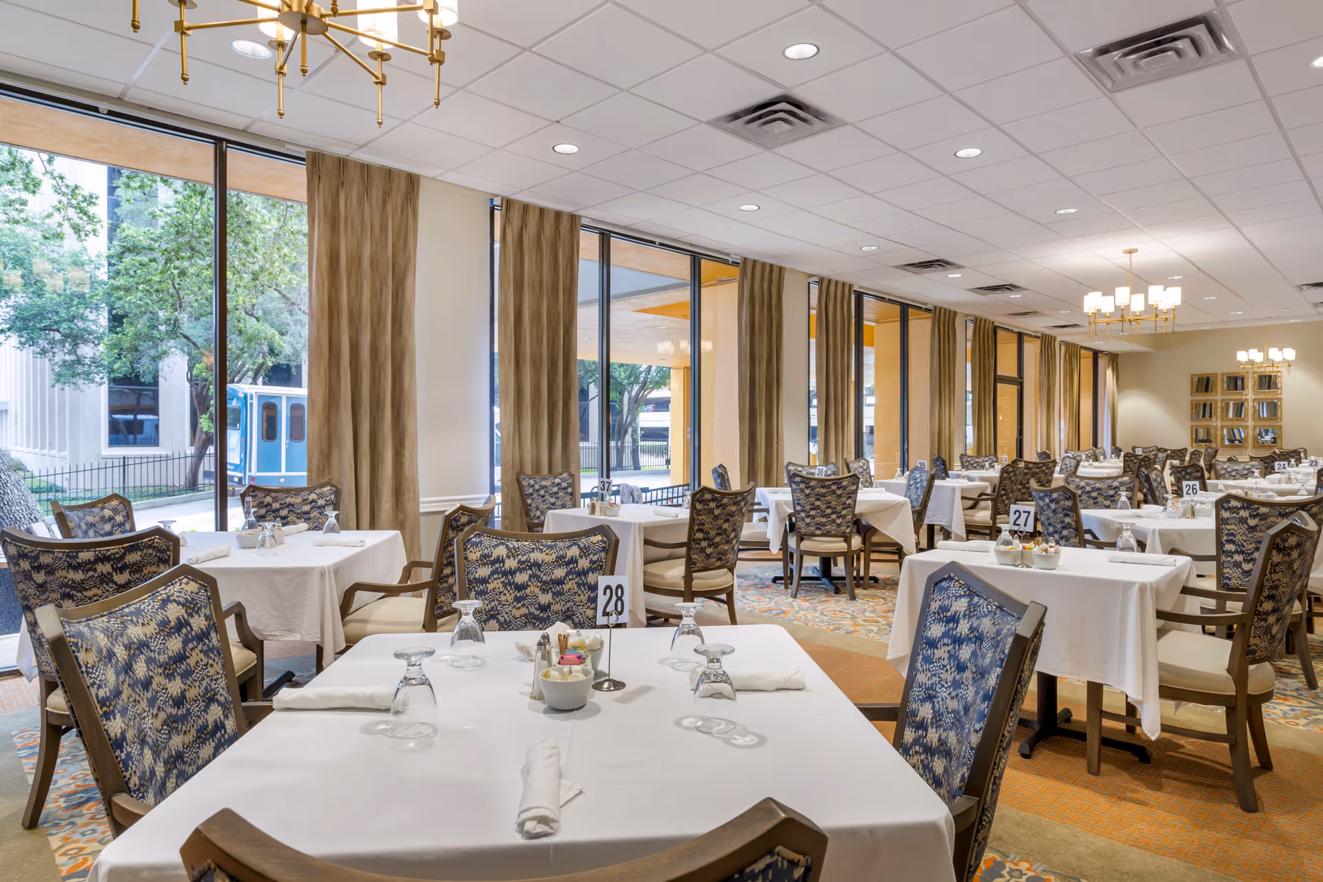 A spacious dining room with multiple tables covered in white tablecloths, each set with upside-down glasses, napkins, and small condiment bowls. The chairs have patterned upholstery in blue and beige tones. Large windows with beige curtains line one side of the room, allowing natural light to fill the space. The ceiling has recessed lighting and modern chandeliers.