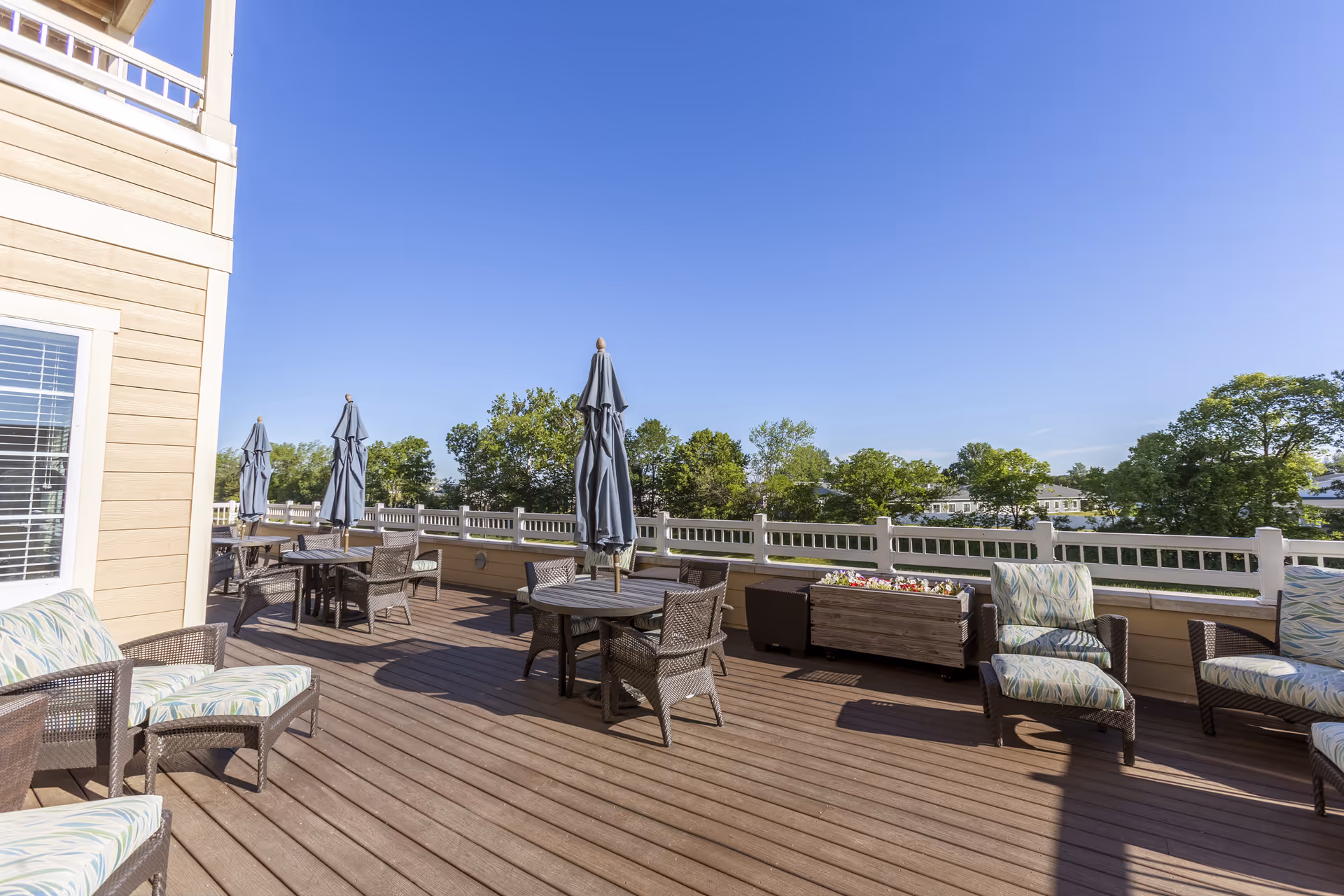 Outdoor patio area with wooden deck flooring, several round tables with chairs, closed umbrellas, cushioned lounge chairs, and planter boxes with flowers. The patio is surrounded by a white railing and overlooks green trees under a clear blue sky.