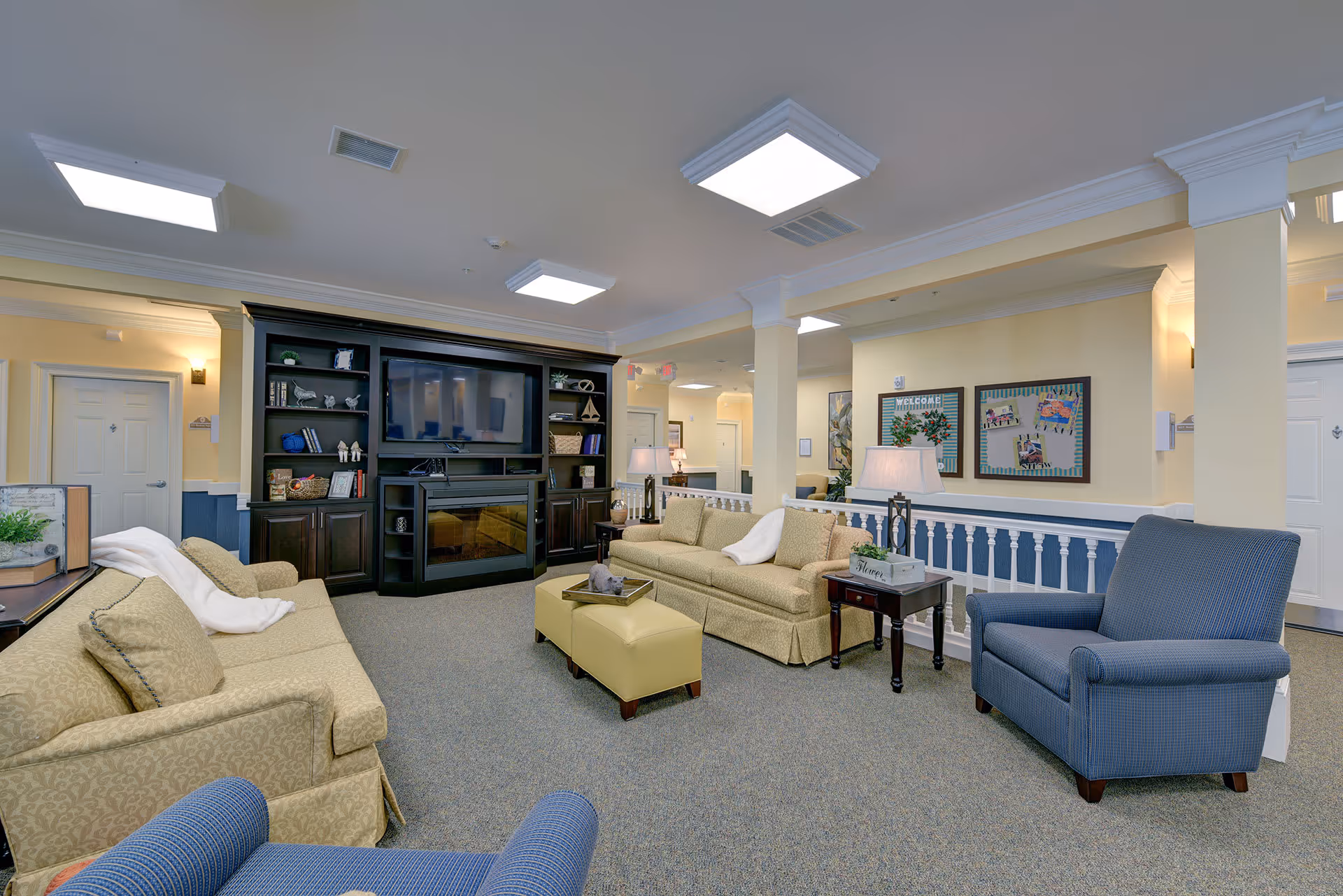 A spacious and well-lit living room area in The Addison of Bluffton featuring two beige sofas, a blue armchair, a yellow ottoman, and a dark wood entertainment center with a large TV and decorative items. The room has beige walls, carpeted floor, and ceiling lights, with framed bulletin boards on the wall and a small side table with a lamp.