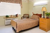 A single bed with a brown patterned bedspread in a small, tidy room. Next to the bed is a wooden nightstand with a lamp and a potted plant. There is a chair with a cushion near the window, which has closed blinds and a valance. The walls are light-colored with a floral border near the ceiling.