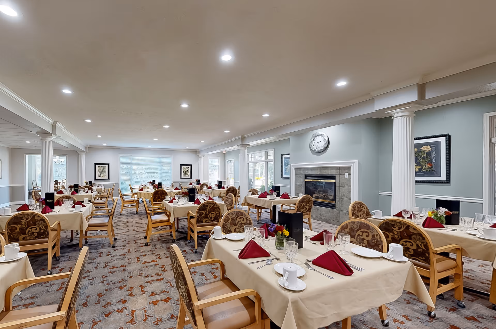 A spacious dining room in an assisted living facility with multiple tables covered in beige tablecloths, each set with white plates, cups, glassware, silverware, and folded red napkins. The room features patterned chairs, a carpeted floor, a fireplace with a clock above it, framed artwork on the walls, and large windows allowing natural light to enter.