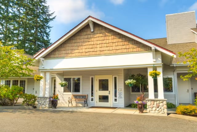 Front exterior view of a single-story building with a covered entrance supported by white columns and stone bases. There are hanging flower baskets and potted plants near the entrance, a bench to the left of the door, and trees and shrubs surrounding the building under a partly cloudy sky.