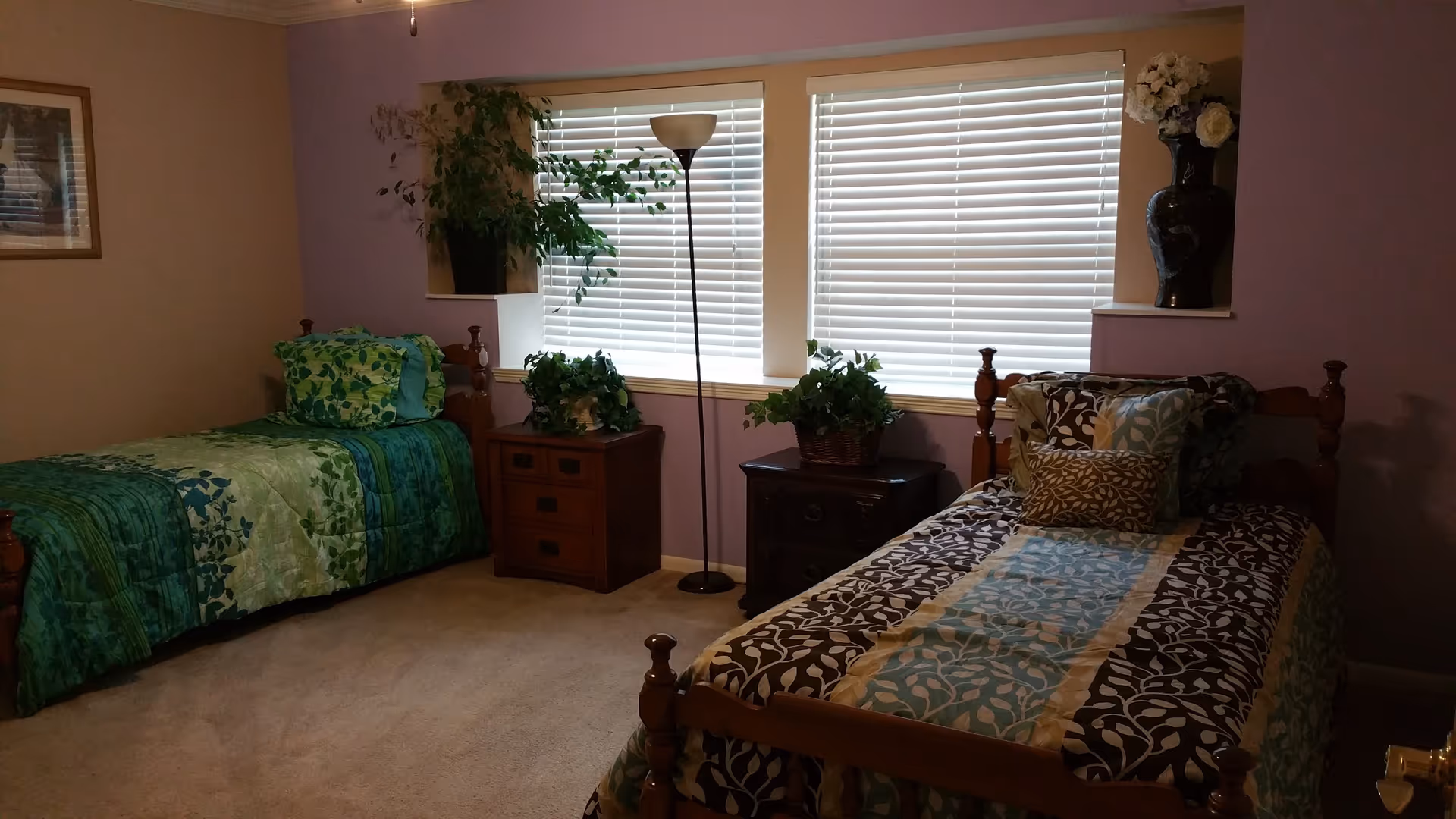 A bedroom with two twin beds, each with patterned bedspreads in green and blue tones. Between the beds are two wooden nightstands with potted plants on top. A tall floor lamp stands in front of two windows with white blinds. The walls are painted in a combination of light purple and beige, and there is a framed picture on the left wall and a vase with flowers on a shelf on the right.
