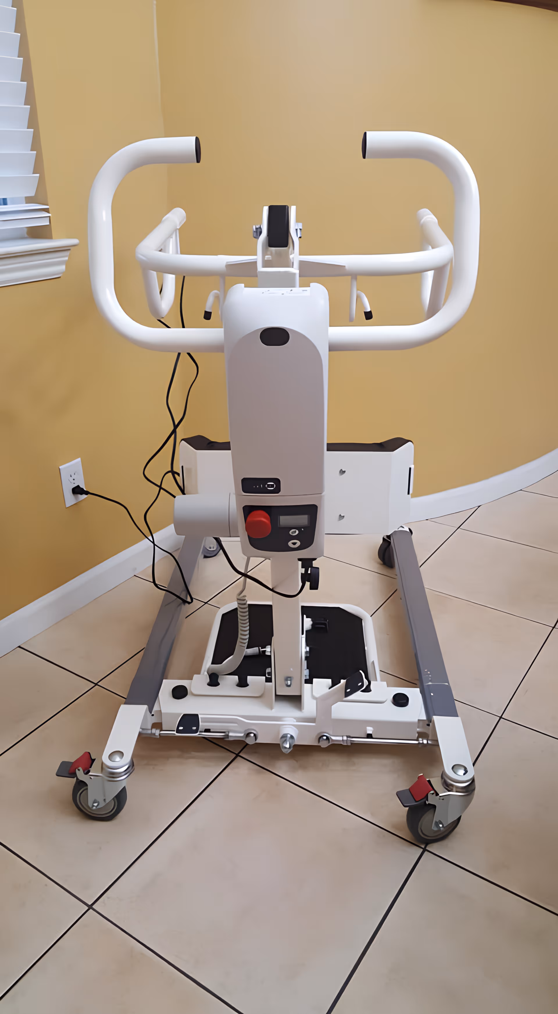 A white medical patient lift device with handles and wheels positioned on a tiled floor against a beige wall with a window partially visible on the left.
