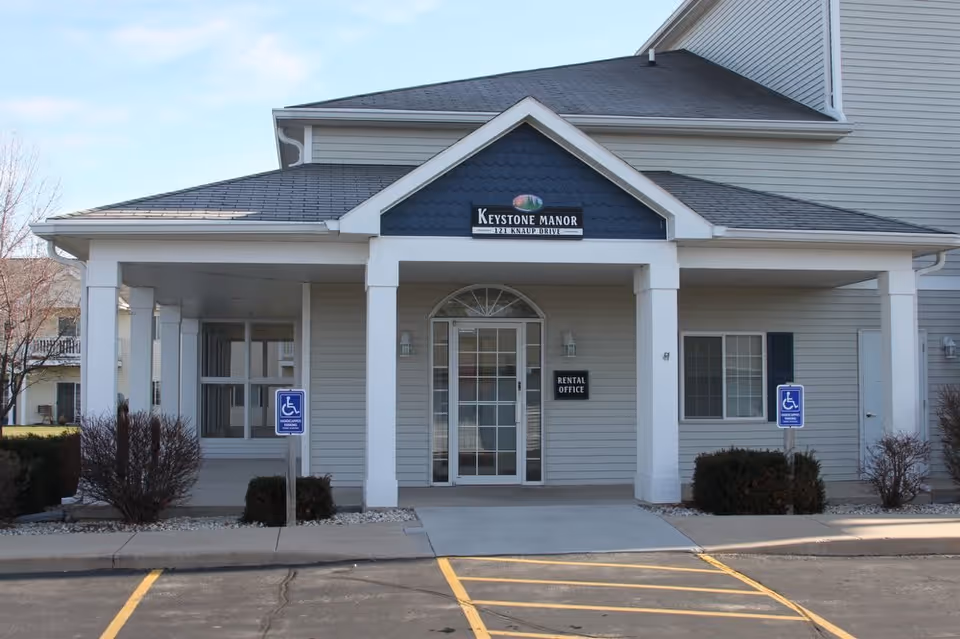 Exterior view of the Keystone Manor Apartments rental office building with a covered entrance, glass door, and two handicap parking signs in front.