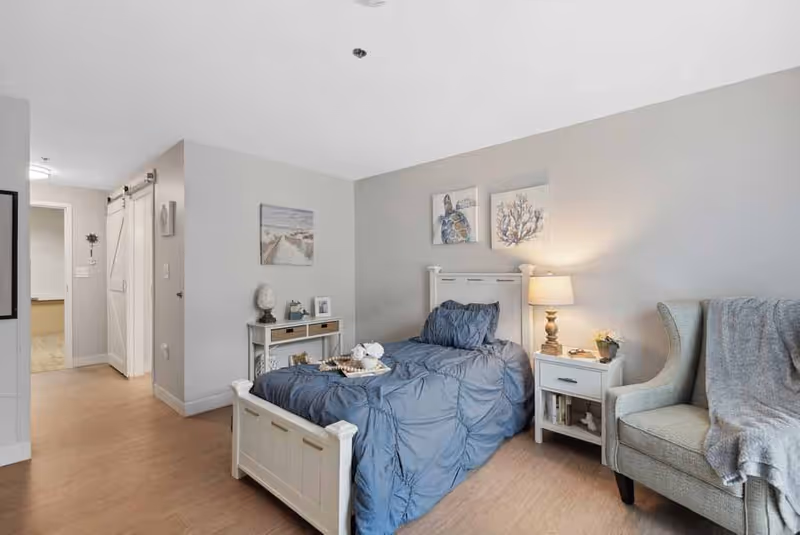 A cozy bedroom with a single bed dressed in blue bedding, a white nightstand with a lamp and small plant, a comfortable armchair with a gray throw blanket, and wall art featuring sea turtles and coral. The room has light gray walls and wood flooring, with a small console table and a barn-style sliding door visible in the background.