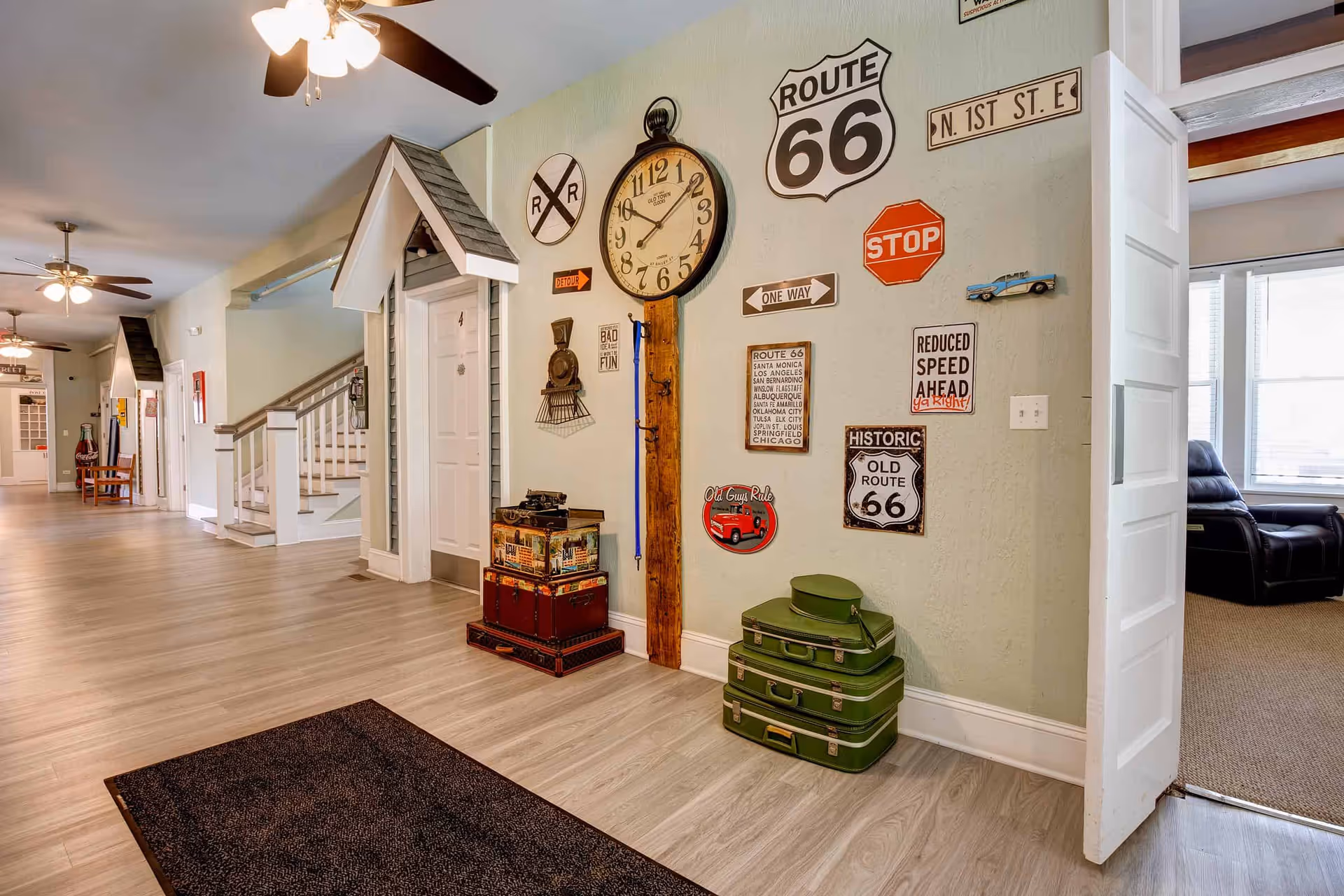 Bright interior hallway decorated with Route 66 signs, a large wall clock, stacked vintage suitcases, and ceiling fans.