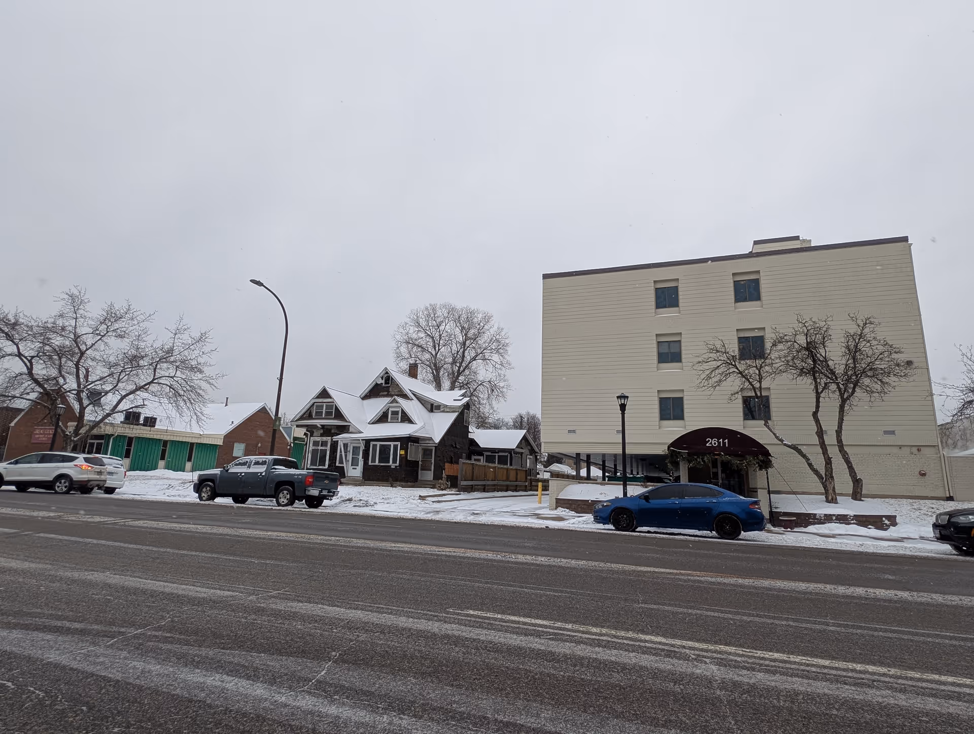 Snowy street view showing a four-story building with an awning marked 2611, adjacent houses, parked cars, and leafless trees under an overcast sky.
